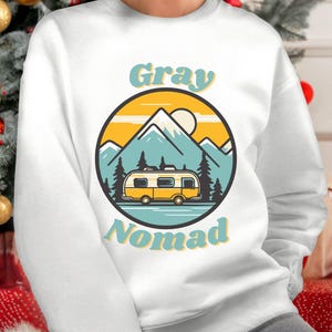 K&ouml;nnte beinhalten: Wei&szlig;er Pullover mit einem Retro-Grafikdesign. Das Design zeigt einen gelben Wohnwagen vor einer Berglandschaft mit Sonnenuntergang. Der Text "Gray Nomad" ist in einer Retro-Schriftart geschrieben.