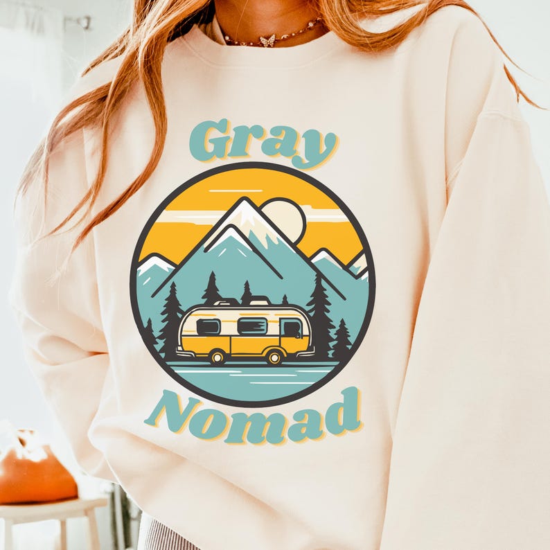 K&ouml;nnte beinhalten: Ein cremefarbener Pullover mit einer Retro-Grafik eines gelben Wohnmobils vor einer Bergkette mit einer Sonne und dem Text "Gray Nomad".