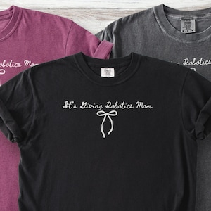 May include: Three t-shirts in shades of black, burgundy, and gray. The black t-shirt features the text "It's Giving Robotics Mom" with a bow graphic. The other shirts have partial text visible. The shirts are made of a soft material.