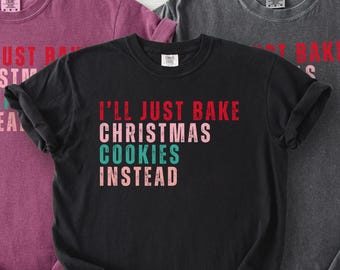 May include: Three t-shirts in shades of burgundy, black, and charcoal gray. The black t-shirt features the text "I'll Just Bake Christmas Cookies Instead" in red, pink, and teal. The other shirts have partial text visible.