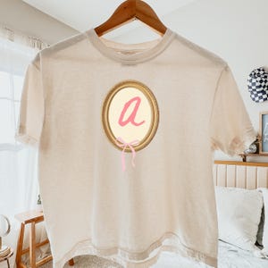 May include: A cream-colored t-shirt with a gold-framed oval design. Inside the oval is a pink letter "a" and a pink bow. The shirt is hanging on a wooden hanger.