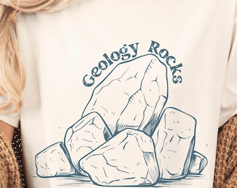May include: Cream-colored t-shirt featuring a blue illustration of several large rocks. The words "Geology Rocks" are arched above the rocks in a matching blue font. The shirt is made of a soft material.