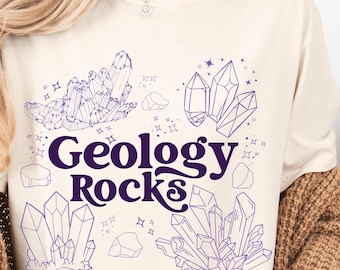 May include: Cream-colored t-shirt featuring a purple graphic design. The design includes illustrations of crystals, rocks, and the text "Geology Rocks" in a stylized font. The shirt is made of a soft material.