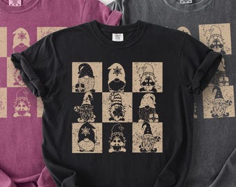 May include: Three t-shirts in shades of burgundy, black, and charcoal gray. The black t-shirt features a grid of nine gnome illustrations in beige and black. The gnomes wear hats and hold gifts. The t-shirts are made of a soft material.