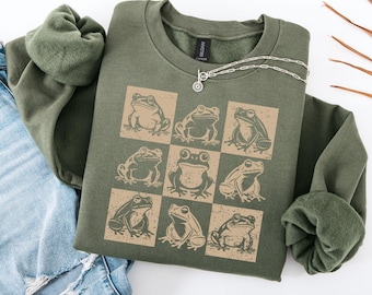 May include: An olive green sweatshirt featuring a grid of nine frog illustrations in tan squares. The sweatshirt has a crew neck and long sleeves. A silver necklace with a small pendant is draped on the shirt. The sweatshirt is folded on top of a pair of blue jeans.