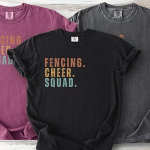 May include: Three short-sleeved t-shirts in shades of black, purple, and gray. The black shirt features the text "FENCING. CHEER. SQUAD." in orange, yellow, and blue. The shirts are made of a soft material.