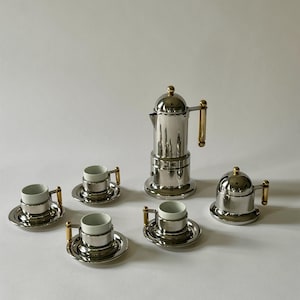 May include: A silver espresso maker set with six cups and saucers. The set includes a coffee pot with a gold handle and lid, and a small lid. The cups have white interiors and gold handles. The saucers are silver.