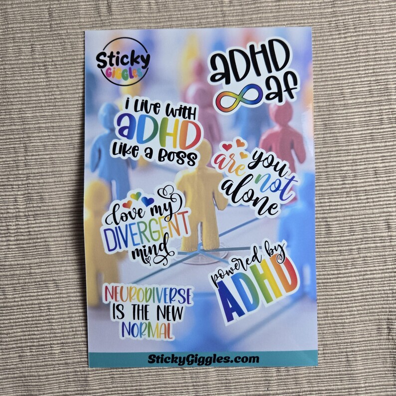 Puede incluir: Una hoja de pegatinas con varias pegatinas coloridas. Las pegatinas tienen frases como "I live with ADHD like a boss" y "Neurodiverse is the new normal". El fondo presenta figuras borrosas.