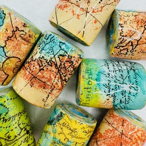 May include: A collection of cylindrical beads with a map design. The beads feature a variety of colors, including yellow, orange, blue, and green. The map design includes lines and text, creating a vintage aesthetic.