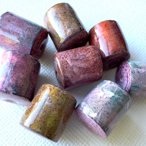 May include: A collection of cylindrical beads in various colors, including shades of pink, brown, and green. The beads have a glossy finish and appear to be made of a natural material. The beads are arranged on a white surface.