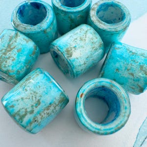May include: A collection of seven cylindrical beads featuring a marbled pattern in shades of bright turquoise, sky blue, and white with subtle metallic gold flecks throughout. Each bead has a smooth, glossy finish and a hollow center opening.
