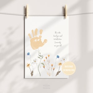 以下が含まれることがあります： 明るいオレンジ色の手形が特徴のプリントで、下部に花のイラストが描かれています。テキストには「私がまだ1歳だったときにあなたが握る必要がある手です」と書かれています。プリントは木製のクリップで保持されています。