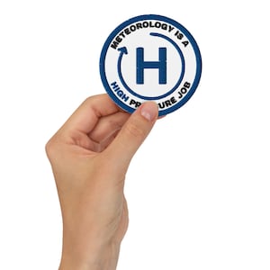 Op de afbeelding: Een ronde, wit en blauwe patch met de tekst "METEOROLOGY IS A HIGH PRESSURE JOB" rondom een grote blauwe "H" en een pijl. De patch wordt door een hand tegen een witte achtergrond gehouden.
