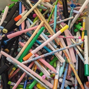 Könnte beinhalten: Ein großer Behälter gefüllt mit einer Vielzahl von bunten Stiften und Markern. Die Stifte sind in verschiedenen Farben, darunter rosa, blau, grün, gelb, orange und schwarz. Einige der Stifte haben dekorative Designs, wie Herzen, Sterne und Blumen.