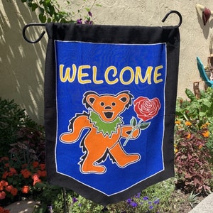 Puede incluir: Una bandera de jardín de bienvenida azul y negra con un oso de dibujos animados sosteniendo una rosa roja. El oso es naranja y lleva un collar verde. El texto "WELCOME" está en letras amarillas.