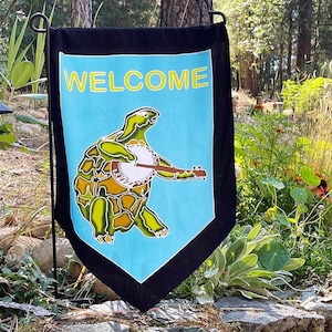 Op de afbeelding: Een decoratieve tuinvaandel met een zwarte rand en een lichtblauw midden. De vlag toont het woord "WELCOME" in gele letters boven een groene schildpad die banjo speelt. De schildpad heeft gele en bruine accenten.