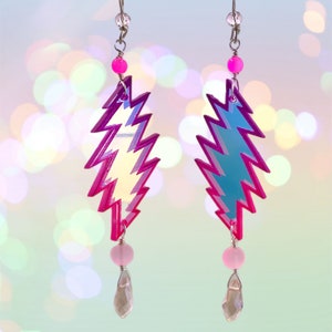 May include: A pair of iridescent lightning bolt earrings with pink beads and crystal drops. The earrings are made of acrylic and have a holographic finish.
