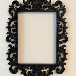 May include: Ornate black picture frame with a rectangular opening. The frame features intricate, swirling floral and leaf designs. The frame is made of a dark, matte material and is mounted on a light-colored wall.