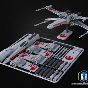 May include: A model kit of a Star Wars X-wing fighter, with a completed model on a stand and unassembled parts in a tray. The fighter is gray with red accents. The stand has a red Rebel Alliance symbol.