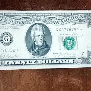 Pode incluir: Uma nota de vinte dólares dos Estados Unidos com um retrato de Andrew Jackson ao centro, o texto UNITED STATES OF AMERICA, o número 20 nos cantos e o número de série G03776752 com um símbolo de estrela no final da sequência.