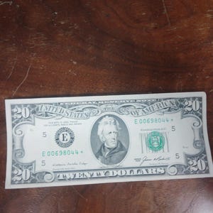 Pode incluir: Uma nota de vinte dólares dos Estados Unidos da América. A nota apresenta um retrato de Andrew Jackson e o texto "Twenty Dollars". A nota é verde e preta com o número de série E 00698044.