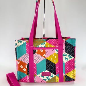 May include: A colorful tote bag with a patchwork design featuring geometric shapes, butterflies, and zebras. The bag has bright pink handles and a matching adjustable shoulder strap. The bag has a zippered pocket.