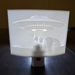 May include: A white night light with a 3D printed image of a UFO hovering over a cow and a figure in a field. The night light has a standard plug-in base.