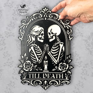 May include: Black and white wall decor featuring two skeletons holding hands, with a candle between them. The phrase "TILL DEATH" is displayed at the bottom. The design is framed with decorative flourishes and roses.
