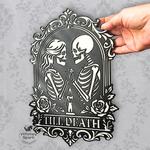 May include: Black and white decorative plaque featuring two skeletons holding hands, framed by an ornate border with roses. The text "TILL DEATH" is displayed at the bottom. The plaque is held by a hand.