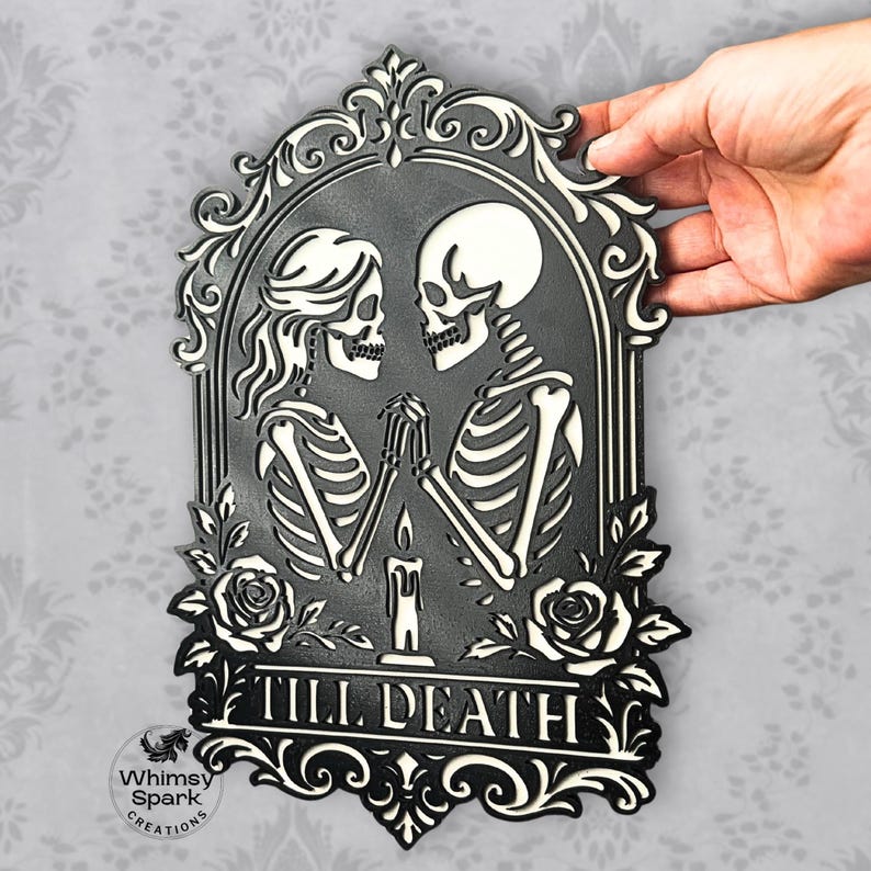 May include: Black and white decorative plaque featuring two skeletons holding hands, framed by an ornate border with roses. The text "TILL DEATH" is displayed at the bottom. The plaque is held by a hand.