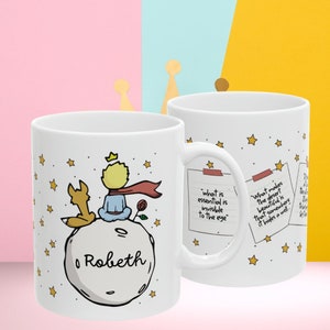 May include: White ceramic mug with a cartoon illustration of The Little Prince sitting on an asteroid with his fox. The mug has a quote from the book, "What is essential is invisible to the eye." The name "Robeth" is written on the asteroid. The mug is on a pink, blue, and yellow background.