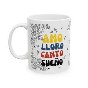 Puede incluir: Taza de cer&aacute;mica blanca con dise&ntilde;os florales negros y el texto "AMO LLORO CANTO SUE&Ntilde;O" en letras de colores. Las letras son de color amarillo, azul, rojo y azul oscuro.