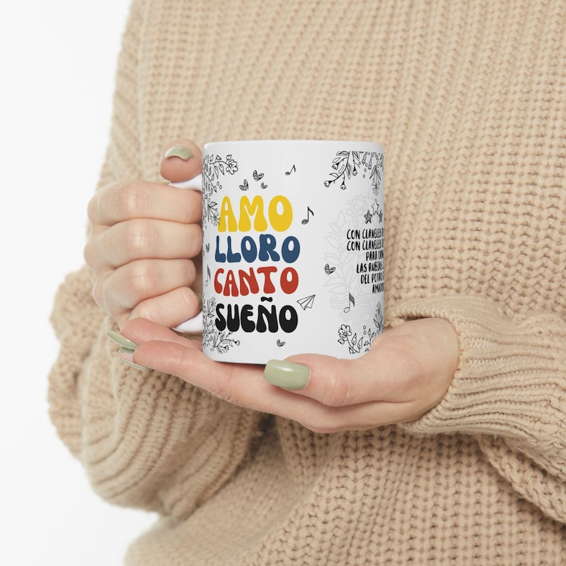 Puede incluir: Taza de cer&aacute;mica blanca con un dise&ntilde;o colorido que presenta las palabras en espa&ntilde;ol "Amo, Lloro, Canto, Sue&ntilde;o" y notas musicales. La taza est&aacute; sostenida por una mano.