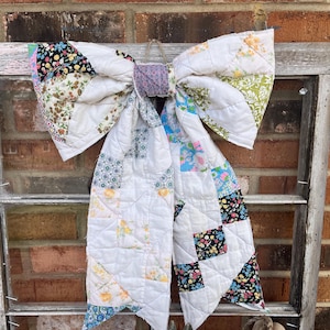 May include: A large, decorative bow made from a patchwork quilt. The bow features a variety of floral and patterned fabrics in white, blue, yellow, and black. It is hanging against a weathered window frame and a brick wall.