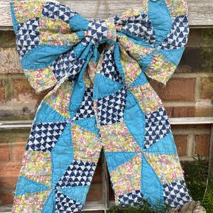May include: A large decorative bow constructed from quilted fabric featuring a patchwork design. The pattern consists of solid bright blue sections interspersed with floral prints in yellow, green, and pink, alongside small dark blue triangle motifs.