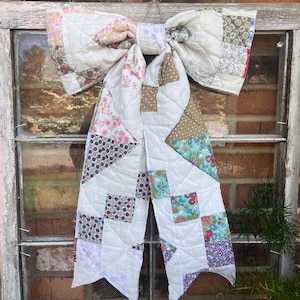 Może przedstawiać: Duża, dekoracyjna kokarda wykonana z patchworkowej kołdry, z różnymi wzorami kwiatowymi i geometrycznymi w odcieniach różu, błękitu i brązu. Kokarda zawieszona jest na naturalnym sznurku, dodając rustykalnego charakteru. Kokarda wisi przed oknem.