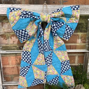 May include: A large decorative bow constructed from quilted fabric featuring a patchwork design. The pattern consists of bright turquoise solid panels, floral sections with pink and green blossoms, and navy blue segments with a white triangle print.