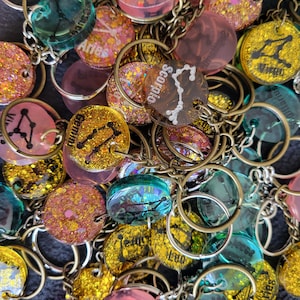 Könnte beinhalten: Nahaufnahme eines Stapels bunter Schlüsselanhänger mit Sternzeichen und Glitzer. Die Schlüsselanhänger sind aus Harz und haben eine Vielzahl von Farben, darunter Pink, Blau, Grün und Gold. Die Schlüsselanhänger sind alle an Metallringen befestigt.
