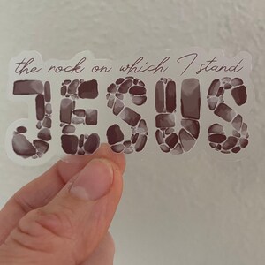 May include: A clear sticker with the words "the rock on which I stand" above the word "JESUS". The word "JESUS" is in large letters, filled with a stone-like pattern in shades of brown and gray.