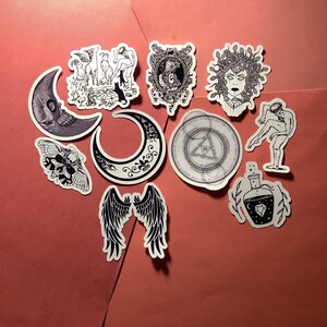 Puede incluir: Una colecci&oacute;n de pegatinas con temas g&oacute;ticos y m&iacute;sticos. Los dise&ntilde;os incluyen una luna calav&eacute;rica, una polilla, una luna creciente, alas de &aacute;ngel, Medusa, una botella de poci&oacute;n y otras ilustraciones en blanco y negro.