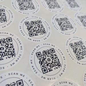 Puede incluir: Una hoja de pegatinas circulares blancas con un código QR negro en el centro. Cada pegatina tiene el texto SCAN ME impreso en un patrón circular alrededor del borde. Algunos códigos QR incluyen pequeños iconos de redes sociales.