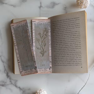 May include: Two vintage-style bookmarks with botanical illustrations and text. One bookmark reads "Happiness" and the other reads "Love." The bookmarks are placed in an open book with aged pages, creating a rustic aesthetic.