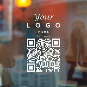 May include: A square white decal featuring the text Your LOGO HERE and EST 2022 above a large QR code. The design is presented on a transparent surface with a blurred background, intended for business branding and digital scanning purposes.
