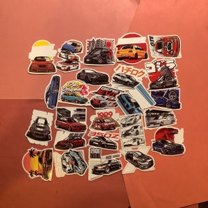 May include: A collection of car-themed stickers featuring various car models in different colors and styles. Some stickers include Japanese text and graphics, while others have text like "Das Original" and "350Z". The stickers are arranged on a pink surface.