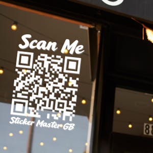 Puede incluir: Un adhesivo de vinilo blanco aplicado sobre una superficie de cristal que muestra un código QR cuadrado. Sobre el código, el texto Scan Me aparece en una fuente blanca y negrita. Debajo del código QR, el texto Sticker Master GB está en cursiva.