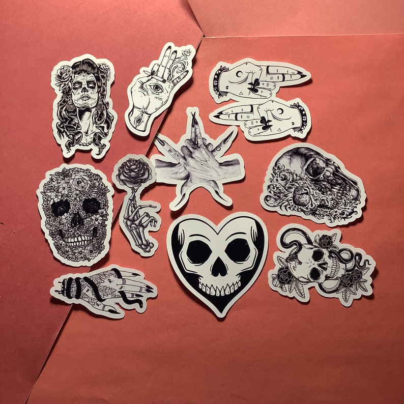 Puede incluir: Una colecci&oacute;n de pegatinas en blanco y negro con dise&ntilde;os g&oacute;ticos y macabros. Las pegatinas incluyen calaveras, manos y una mujer del D&iacute;a de los Muertos. Las pegatinas est&aacute;n sobre un fondo rosa.