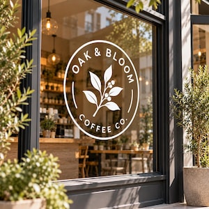 May include: A white circular vinyl decal applied to a glass storefront window. The design features a central botanical illustration of a leafy branch with berries, surrounded by the text OAK & BLOOM COFFEE CO. in a clean, sans-serif font.