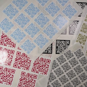 May include: A collection of square sticker sheets featuring multiple QR codes in various colours including light blue, magenta, black, and olive green. Each sheet contains a grid of individual scannable QR code labels with a glossy finish.