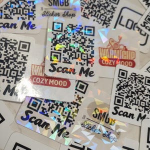 May include: A collection of square holographic stickers featuring black and white QR codes. Some stickers display the text Scan Me, while others show the text SMGB Sticker Shop or Warm Cup Cozy Mood with a small graphic of a cup with arms.