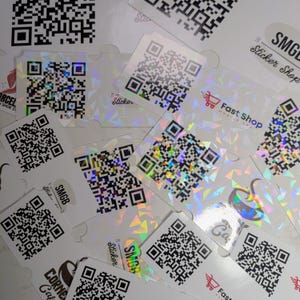 Op de afbeelding: Een verzameling vierkante stickers met zwart-witte QR-codes op een holografische, lichtreflecterende achtergrond. Sommige stickers tonen logo's en tekst zoals Fast Shop, SMGB Sticker Shop en Corner Cafe, verspreid over het oppervlak gerangschikt.